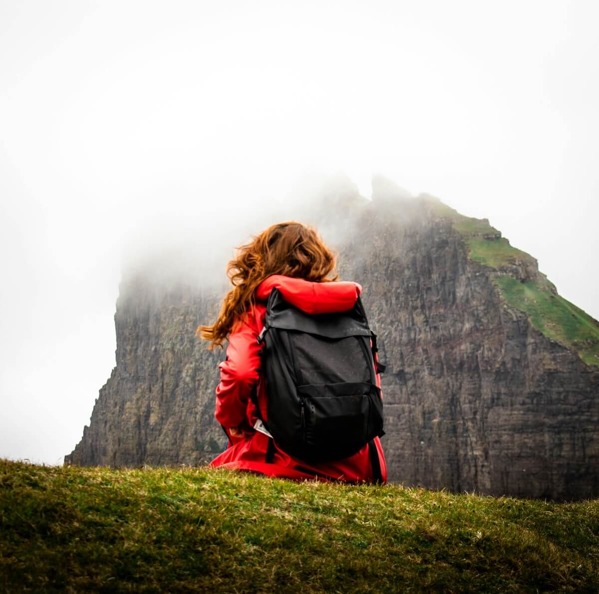 Woman with backpack sitting before fog-covered mountain cliffs