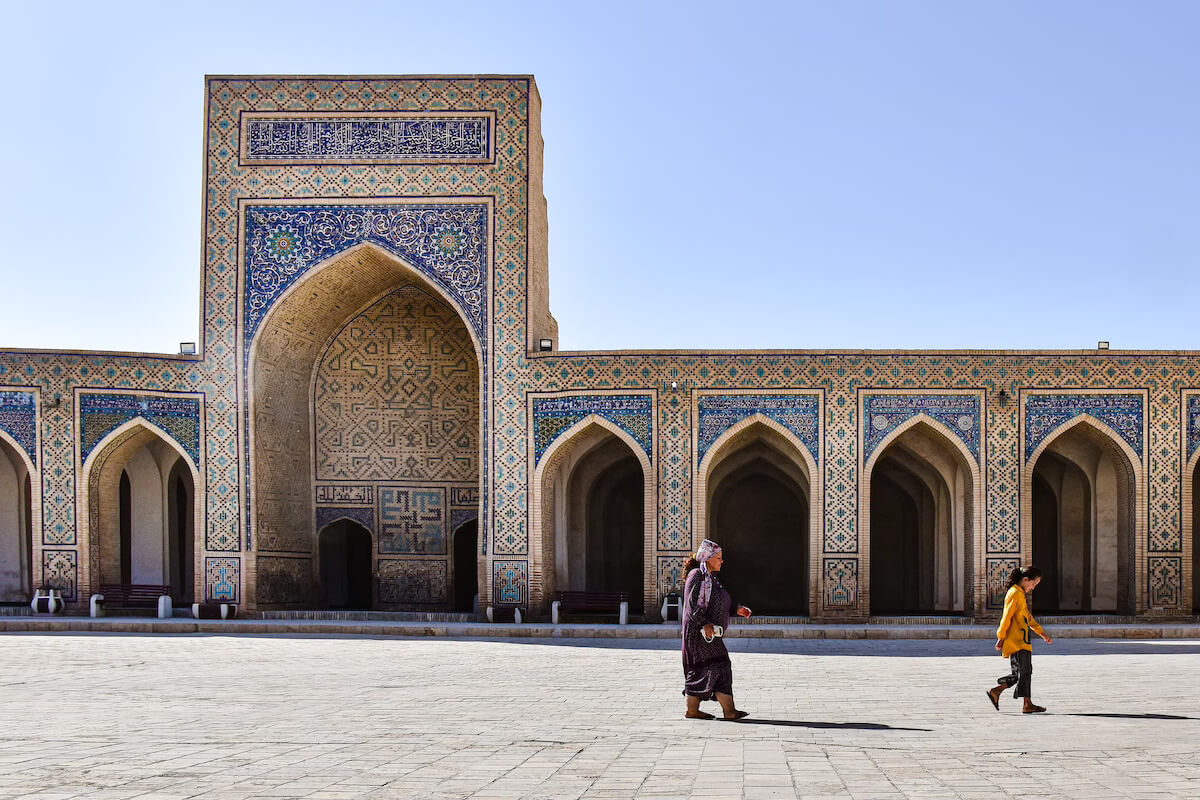 People walking past the tiled entrance of the Kalon Mosque in Bukhara, Uzbekistan, under a clear sky